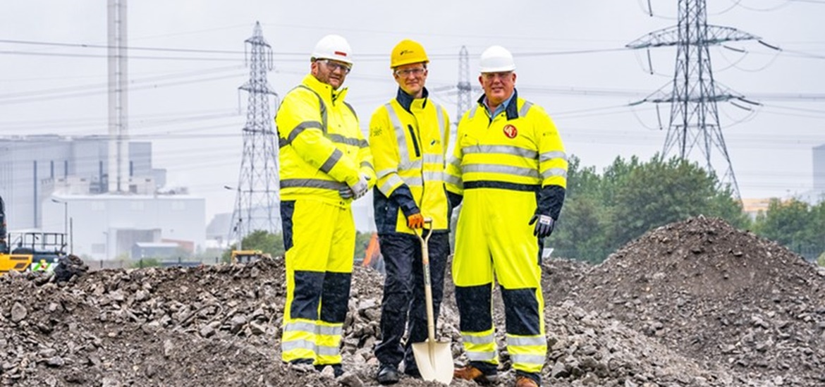 3 SSE Renewables constructor workers on the empty construction site with one power tower in the back, holding a shovel, smiling to the camera